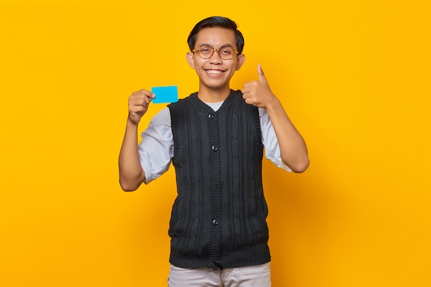 クレジットカードを保持し、黄色の背景に親指を立てるジェスチャーを示す陽気な若いアジア人男性