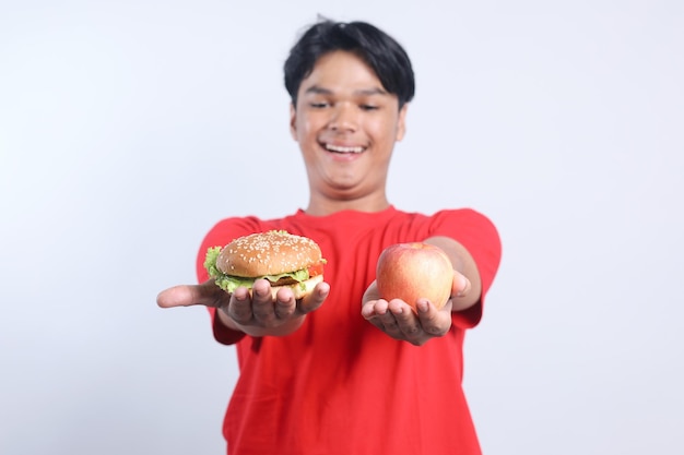 Cheerful young Asian man holding hamburger and apple fruit choosing between healthy or unhealthy foo