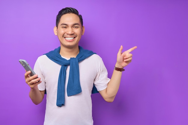 Cheerful young Asian man holding smartphone and pointing aside at copy space isolated over purple background