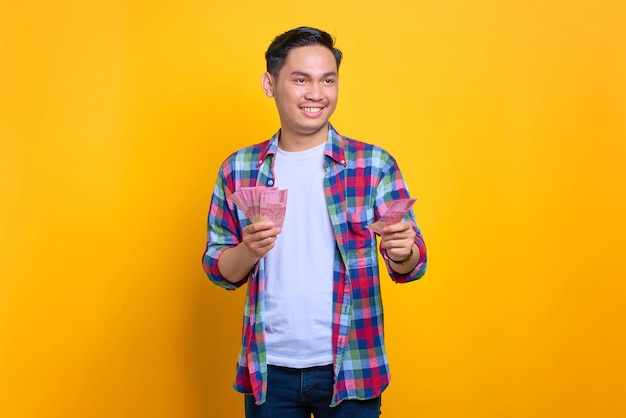 Cheerful young Asian man in plaid shirt giving money banknote to someone isolated on yellow background