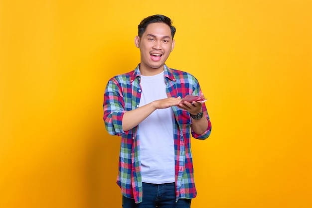 Cheerful young asian man in plaid shirt holding money banknotes and looking at camera isolated on yellow background