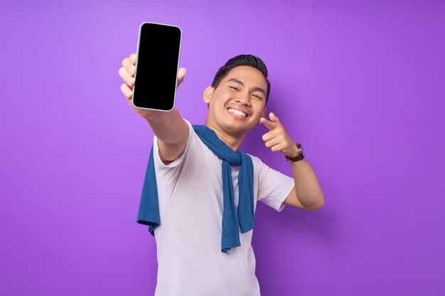 Cheerful young asian man pointing index finger at empty smartphone screen isolated on purple background advertising new mobile app mockup concept