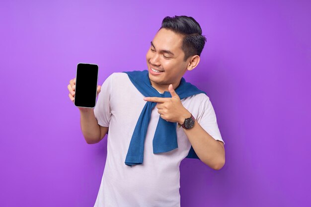 Cheerful young Asian man pointing index finger at empty smartphone screen isolated on purple background Advertising new mobile app mockup concept