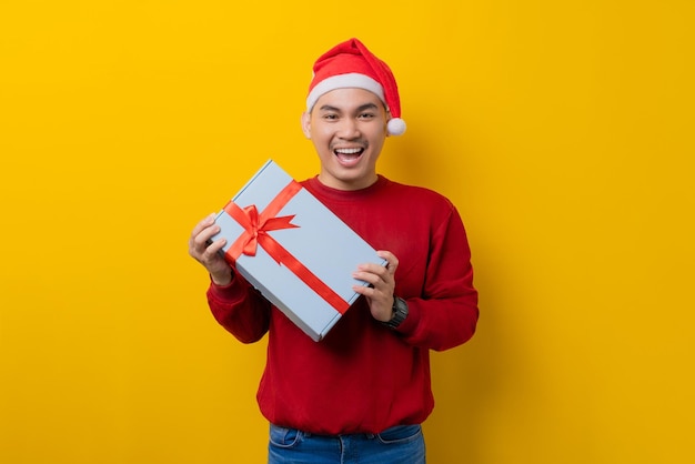 Cheerful young Asian man in Santa hat holding gift box with ribbon on yellow studio background celebration Christmas holiday and New Year concept