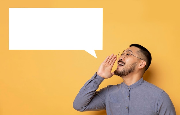 Photo cheerful young asian man shouting at empty speech bubble on yellow background