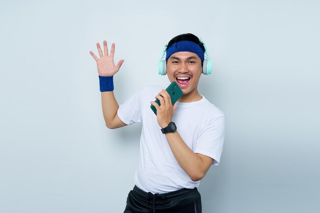 Cheerful young Asian man sporty fitness trainer instructor in blue headband and white tshirt with headphones listening to music sing song in dictaphone on mobile cell phone over white background