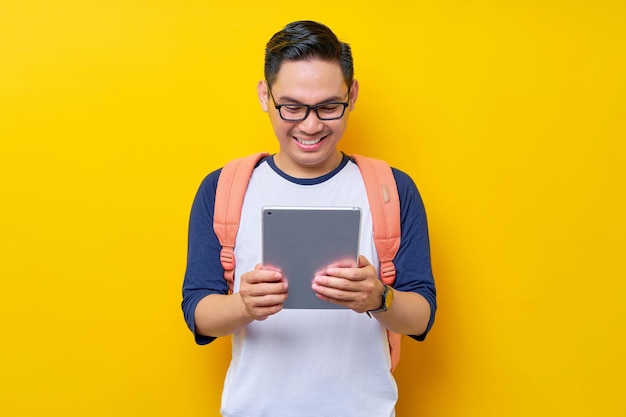 Cheerful young asian man student in casual clothes glasses backpack using digital tablet computer for studying reading ebooks isolated on yellow background high school university college concept