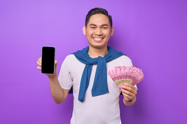 흰색 티셔츠를 입은 쾌활한 젊은 아시아 남자가 빈 화면 휴대폰을 보여주고 보라색 배경에 고립된 지폐를 들고 있습니다.