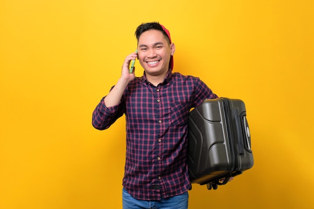 Cheerful young asian man with suitcase talking on smartphone isolated over yellow background tourism and holiday trip concept