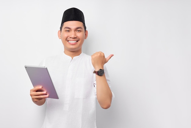 Cheerful young Asian Muslim man holding digital tablet and pointing thumb at copy space isolated on white background People religious Islam lifestyle concept celebration Ramadan and ied Mubarak