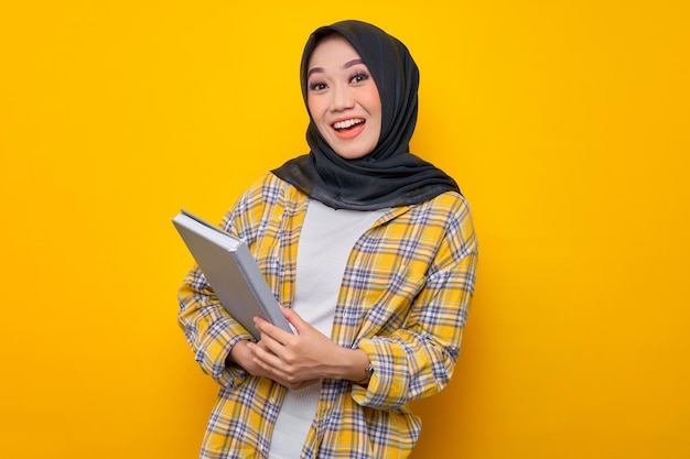 Cheerful young asian muslim student holding book looking at
camera isolated on yellow background back to school in high school
college concept