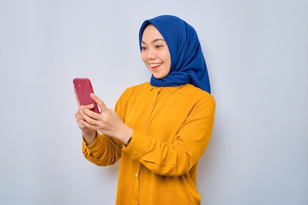 Cheerful young Asian Muslim woman dressed in orange shirt looking at mobile phone screen feels joyful reading good news isolated over white background