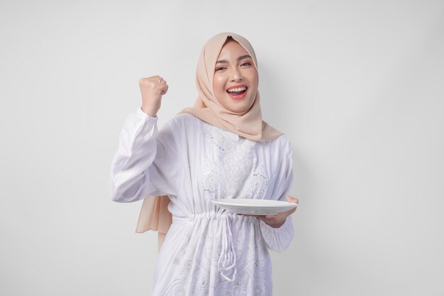 Cheerful young Asian Muslim woman in hijab doing a successful win gesture with clenched fist while presenting an empty plate and copy space over it