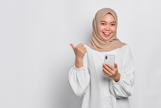 쾌활한 젊은 아시아 이슬람 여성 휴대 전화를 들고 흰색 배경에 고립 된 복사 공간을 가리키는