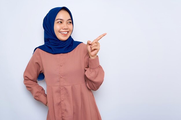 Веселая молодая азиатская мусульманка в розовой рубашке, указывающая пальцем на пространство для копирования, изолированное на белом фоне Концепция религиозного образа жизни людей