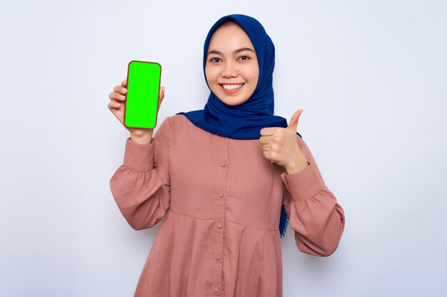 Cheerful young Asian muslim woman in pink shirt showing mobile phone blank screen recommending App and gesturing thumbs up isolated over white background People religious lifestyle concept