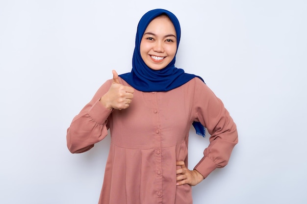 Cheerful young Asian muslim woman in pink shirt showing thumbs up gesture with hand isolated over white background People religious lifestyle concept