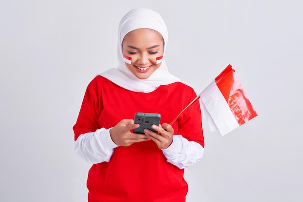흰색 배경에 격리된 8월 17일에 휴대폰을 사용하고 인도네시아 독립 기념일을 축하하는 빨간색 흰색 티셔츠를 입은 쾌활한 젊은 아시아 이슬람 여성