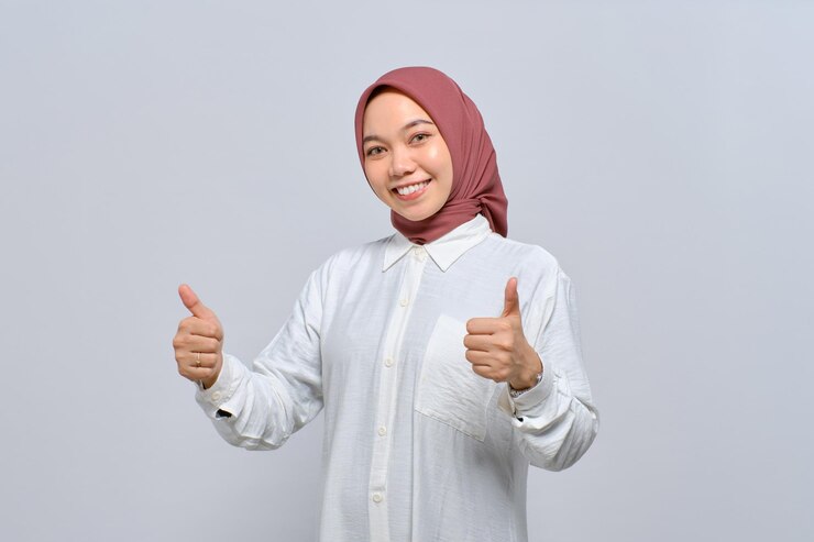 陽気な若いアジアのイスラム教徒の女性親指ジェスチャーを示し、白い背景で隔離されたカメラに笑顔