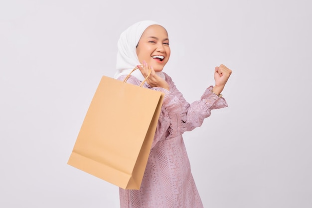 Cheerful young Asian Muslim woman wearing hijab and purple dress holding shopping bag and celebrating victory isolated on white studio background