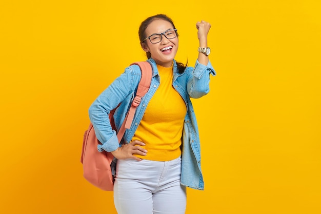 Cheerful young asian student in denim outfit with backpack\
standing doing winning gesture celebrating fist saying yes isolated\
on yellow background education in high school university college\
concept