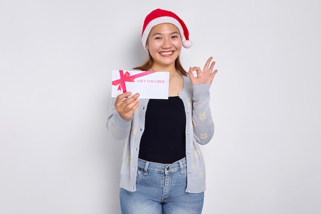 Cheerful young asian woman 20s in hat christmas holding a gift\
certificate coupon voucher card showing okay sign isolated on white\
studio background merry christmas concept