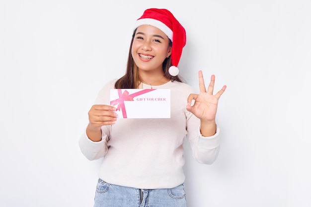 Cheerful young Asian woman 20s in hat Christmas holding a gift certificate coupon voucher card Showing okay sign isolated on white studio background Merry Christmas Concept