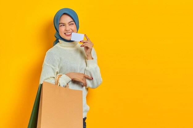 Cheerful young asian woman biting credit card and hold shopping bags on yellow background