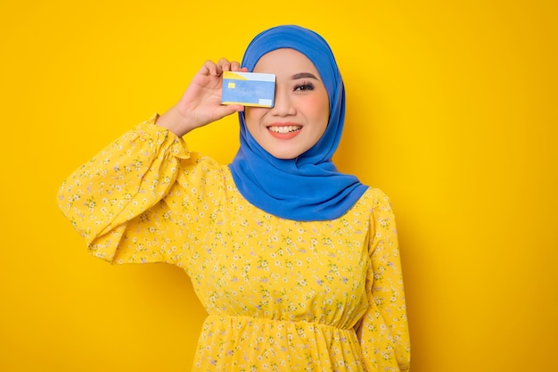 Cheerful young Asian woman in casual dress covering his eye with credit card isolated on yellow background