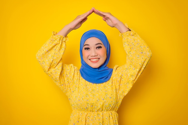 Cheerful young asian woman in casual dress making rooftop home\
gesture with hands above head isolated on yellow background