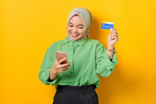 Cheerful young asian woman in a green shirt holding mobile\
phone and credit card rejoicing luck on yellow background