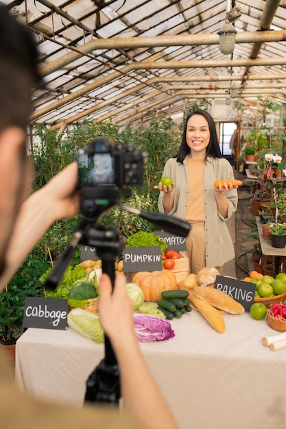 Cheerful young Asian woman holding apple and carrot while sharing healthy recipe with followers shooting video for social media