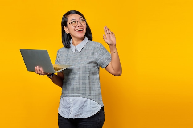 Cheerful young Asian woman holding laptop looking aside and waving on yellow background