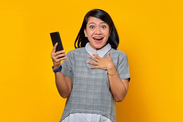 Cheerful young Asian woman holding mobile phone and putting palms on chest over yellow background