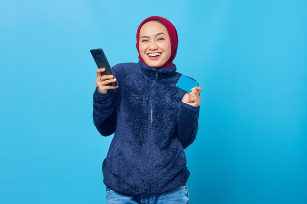 Cheerful young asian woman holding mobile phone and showing credit card on blue background