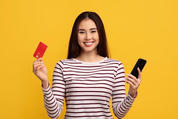 Cheerful young asian woman holding phone and credit card