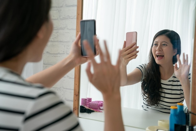 Cheerful young asian woman is sitting at dressing table with smile talking chatting on cellphone video phone call with friend. happy girl waving hands saying hi to family online on internet mobile.