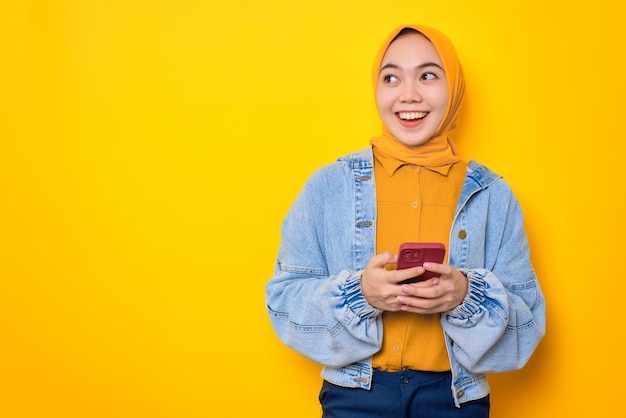 Cheerful young Asian woman in jeans jacket holding mobile phone and looking aside at copy space isolated over yellow background