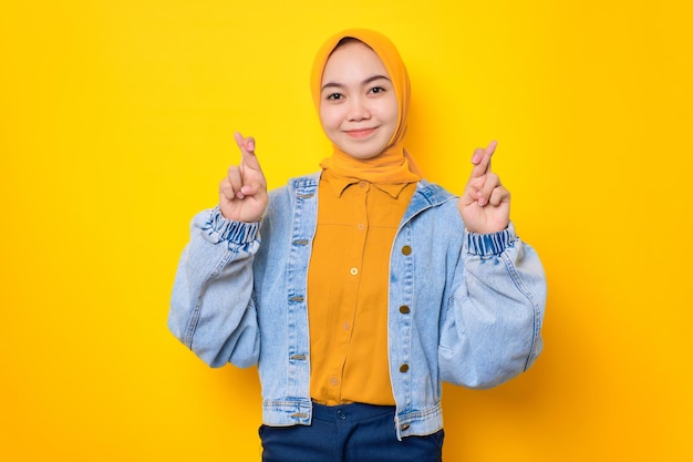 Cheerful young Asian woman in jeans jacket showing peace sign and looking at camera isolated over yellow background