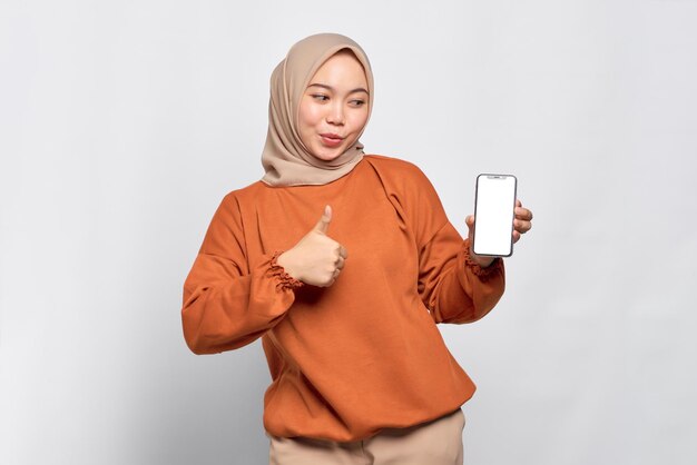 Cheerful young Asian woman in orange shirt showing mobile phone blank screen recommending App and gesturing thumbs up isolated over white background