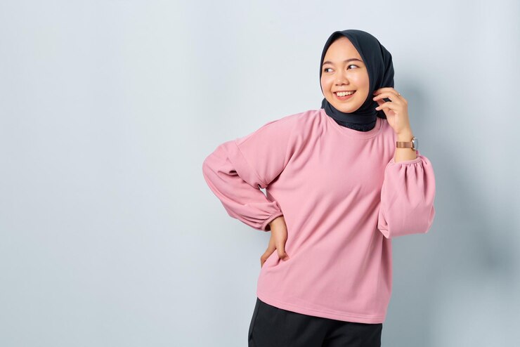 Cheerful young Asian woman in pink shirt holding hand on hip looking away at copy space isolated over white background