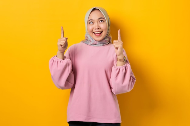 Cheerful young Asian woman in pink shirt pointing finger up at copy space on yellow background