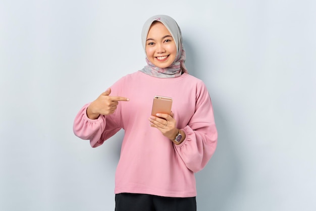 白い背景の上に分離された携帯電話で指を指すピンクのシャツの陽気な若いアジアの女性