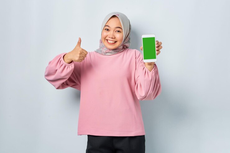 Cheerful young Asian woman in pink shirt showing mobile phone blank screen recommending App and gesturing thumbs up isolated over white background