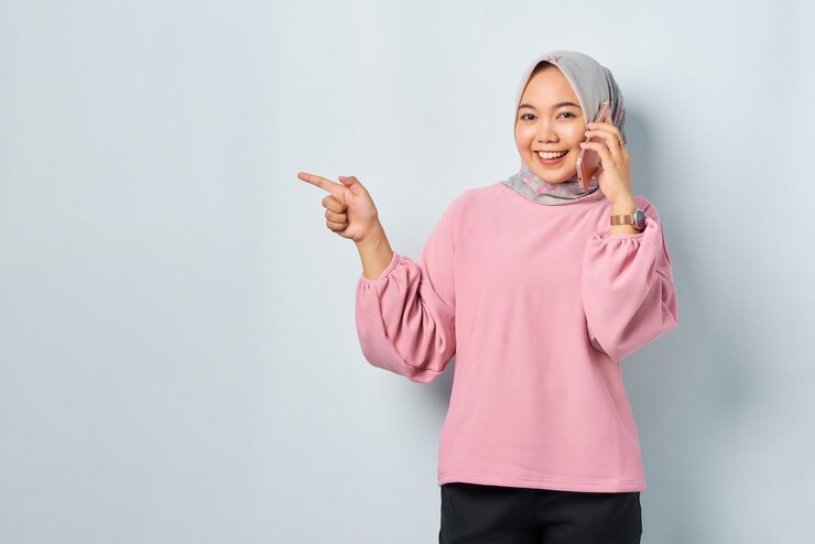 Cheerful young Asian woman in pink shirt talking on mobile phone and pointing finger away at blank space isolated over white background
