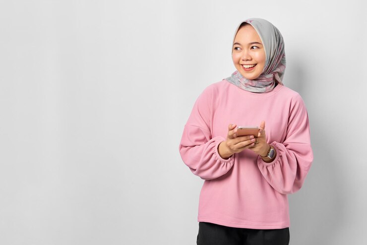 Cheerful young Asian woman in pink shirt using mobile phone and looking away at blank space isolated over white background