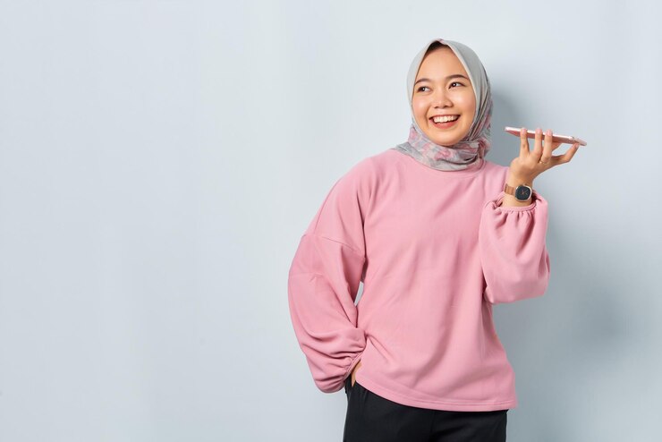 Cheerful young Asian woman in pink shirt using the voice assistant on mobile phone looking away at blank space isolated over white background