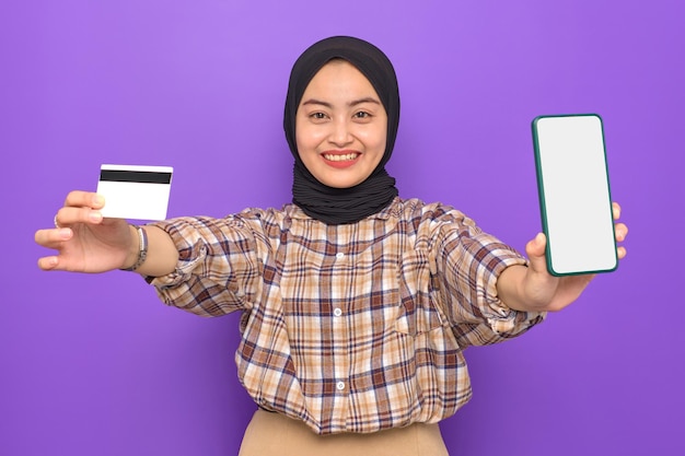 격자 무늬 셔츠를 입은 쾌활한 젊은 아시아 여성이 빈 화면 휴대폰을 보여주고 보라색 배경에 격리된 신용 카드를 들고 있습니다.