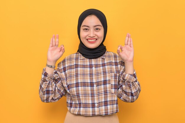 Cheerful young Asian woman in plaid shirt showing okay gesture demonstrates symbol of approval isolated on yellow background