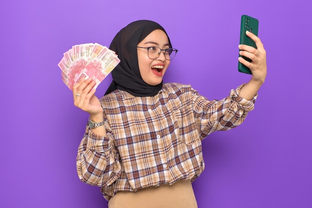 Cheerful young asian woman in plaid shirt using a mobile phone\
and holding money banknotes isolated on purple background